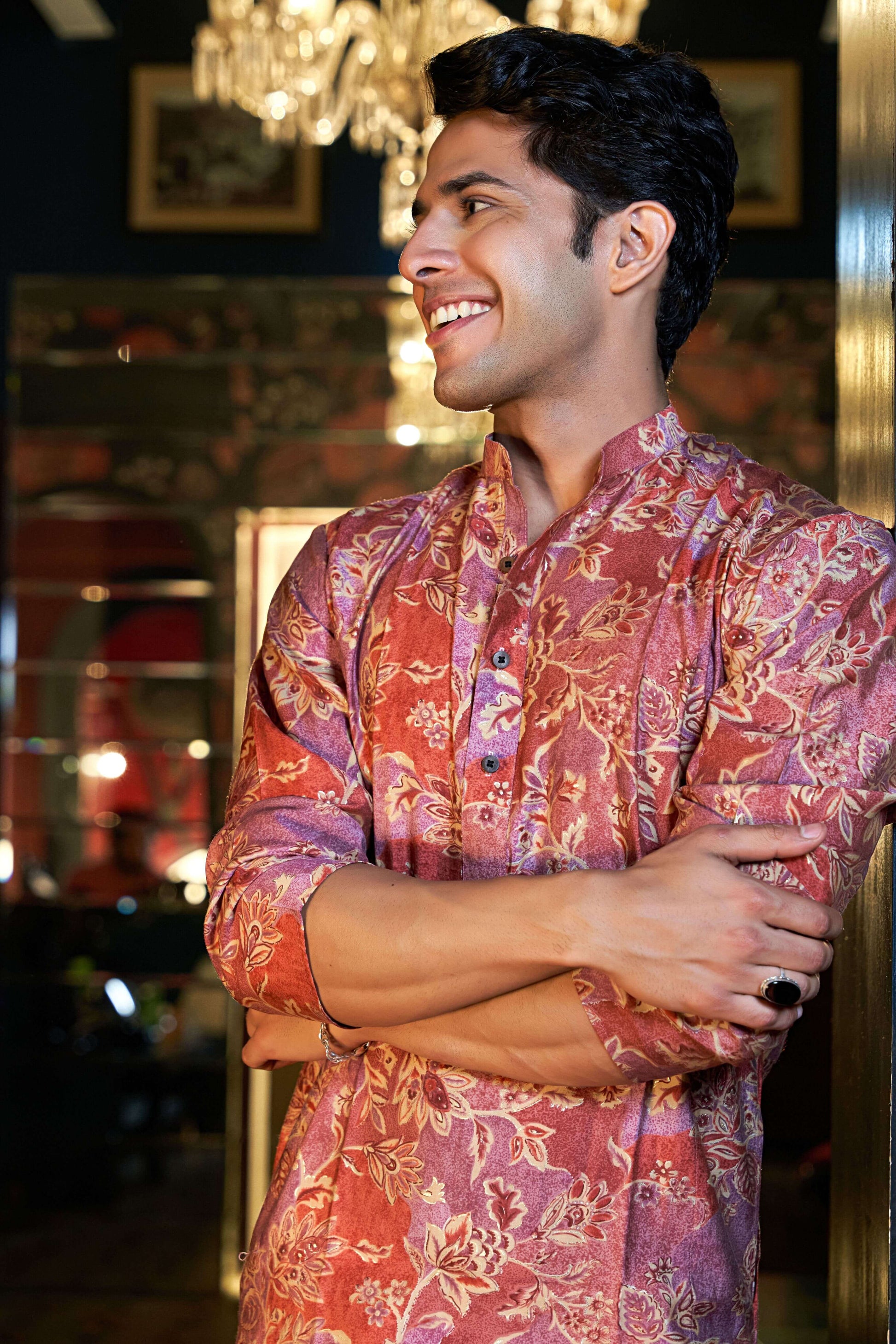 A person wearing a red floral patchwork kurta stands in an indoor setting with a chandelier and framed pictures in the background. The person has their arms crossed, and their face is blurred out, highlighting the detailed design of the traditional attire.