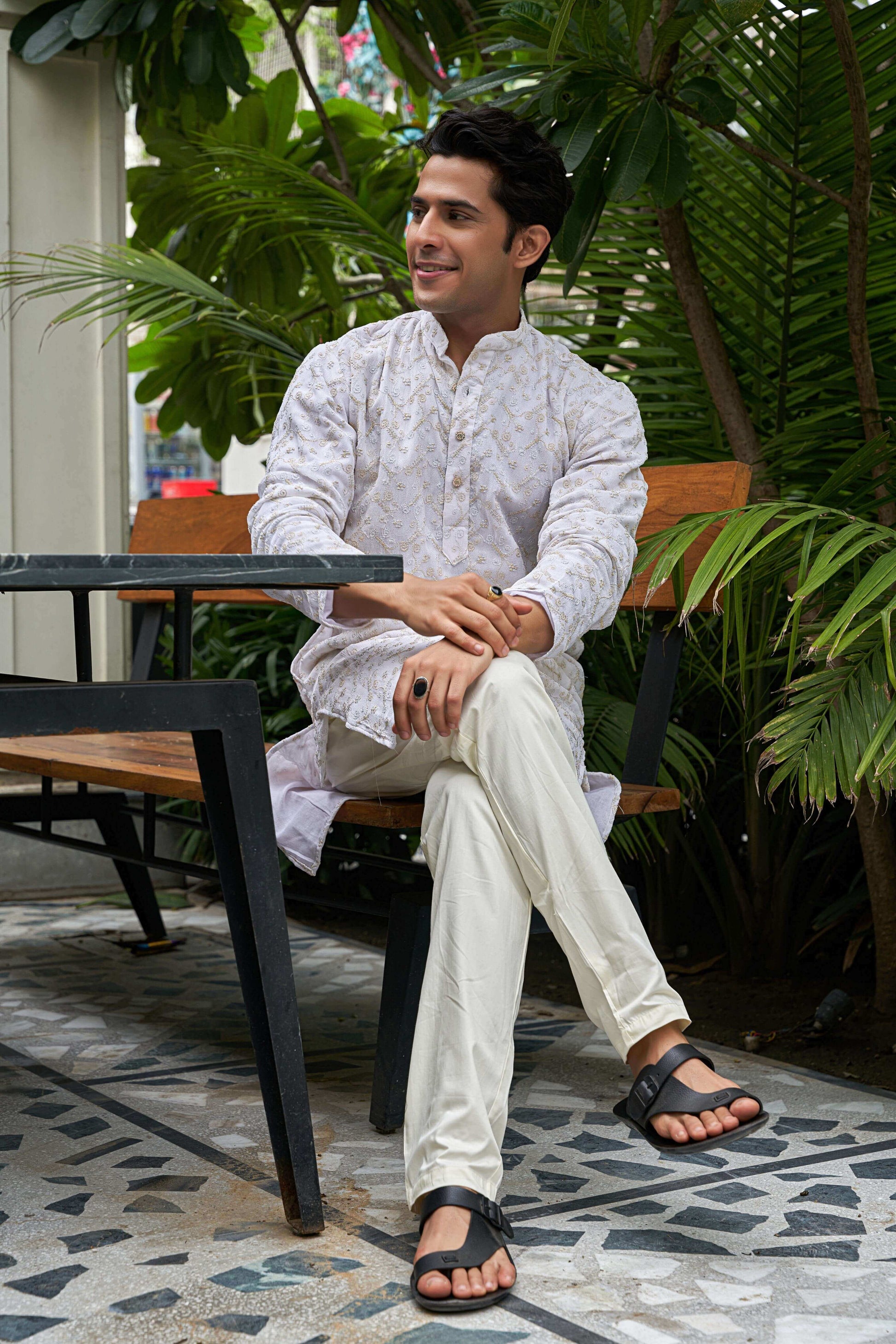 A man in a white zari work kurta sits on a bench.