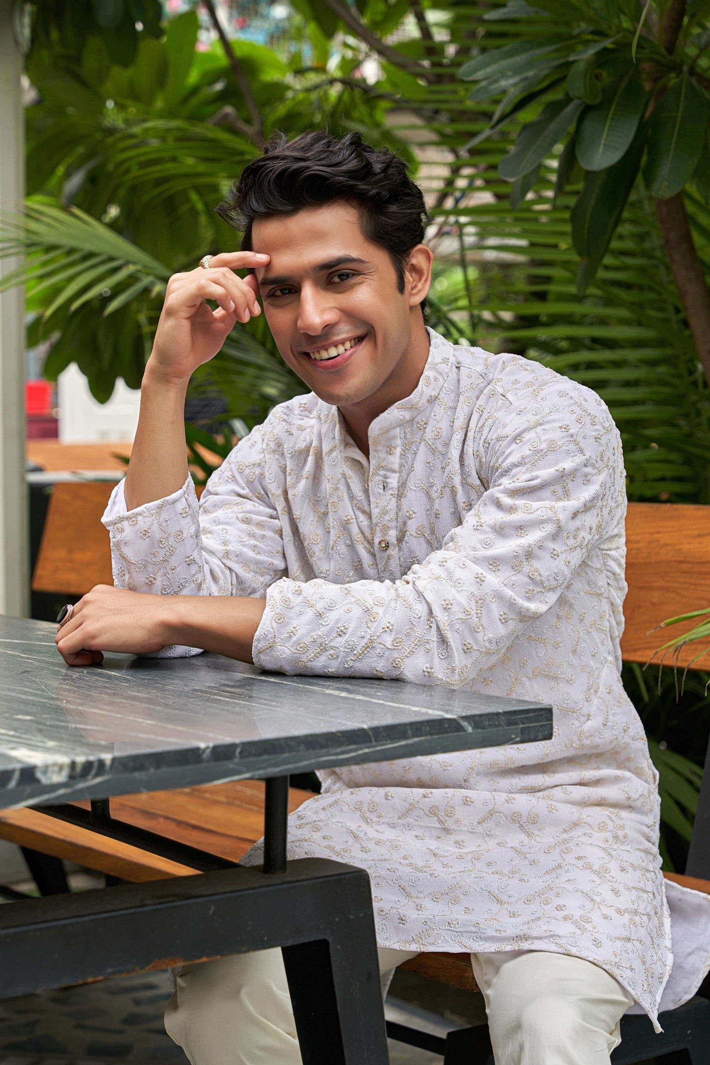 Indian man wearing a white zari work long kurta. sitting on a table.