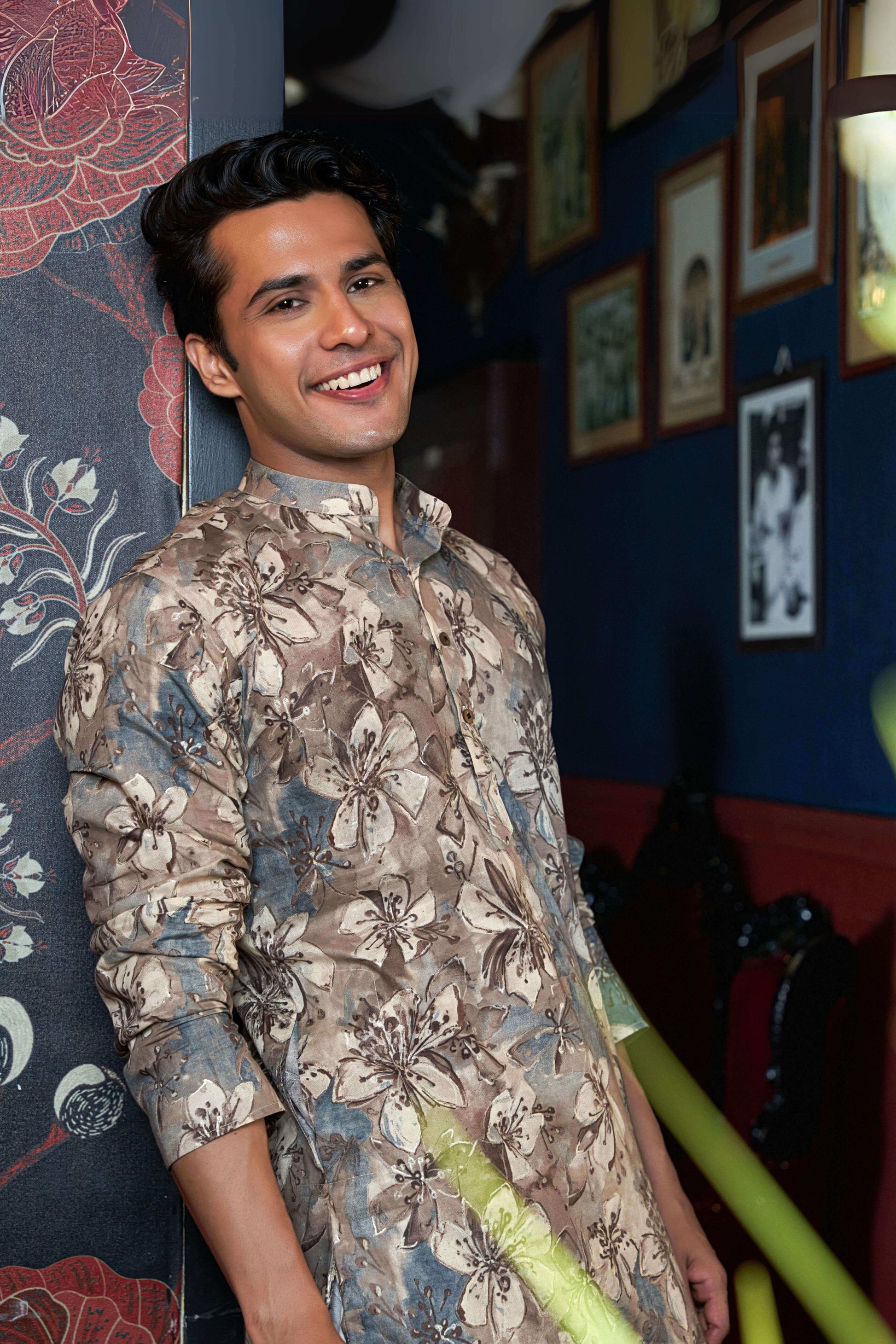 A man wearing a floral printed shirt stands against a textured wall, showcasing a vibrant and casual style.