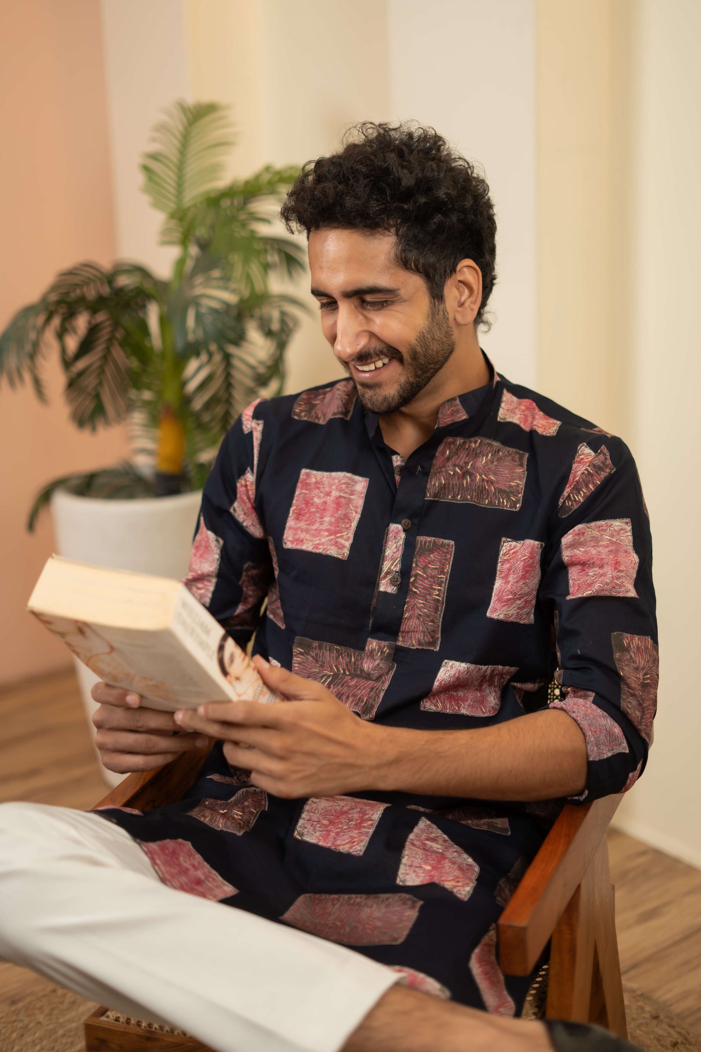 A man sitting on a wooden chair indoors, smiling while reading a book. He is wearing a navy blue kurta with red and beige patchwork patterns and white pants. 