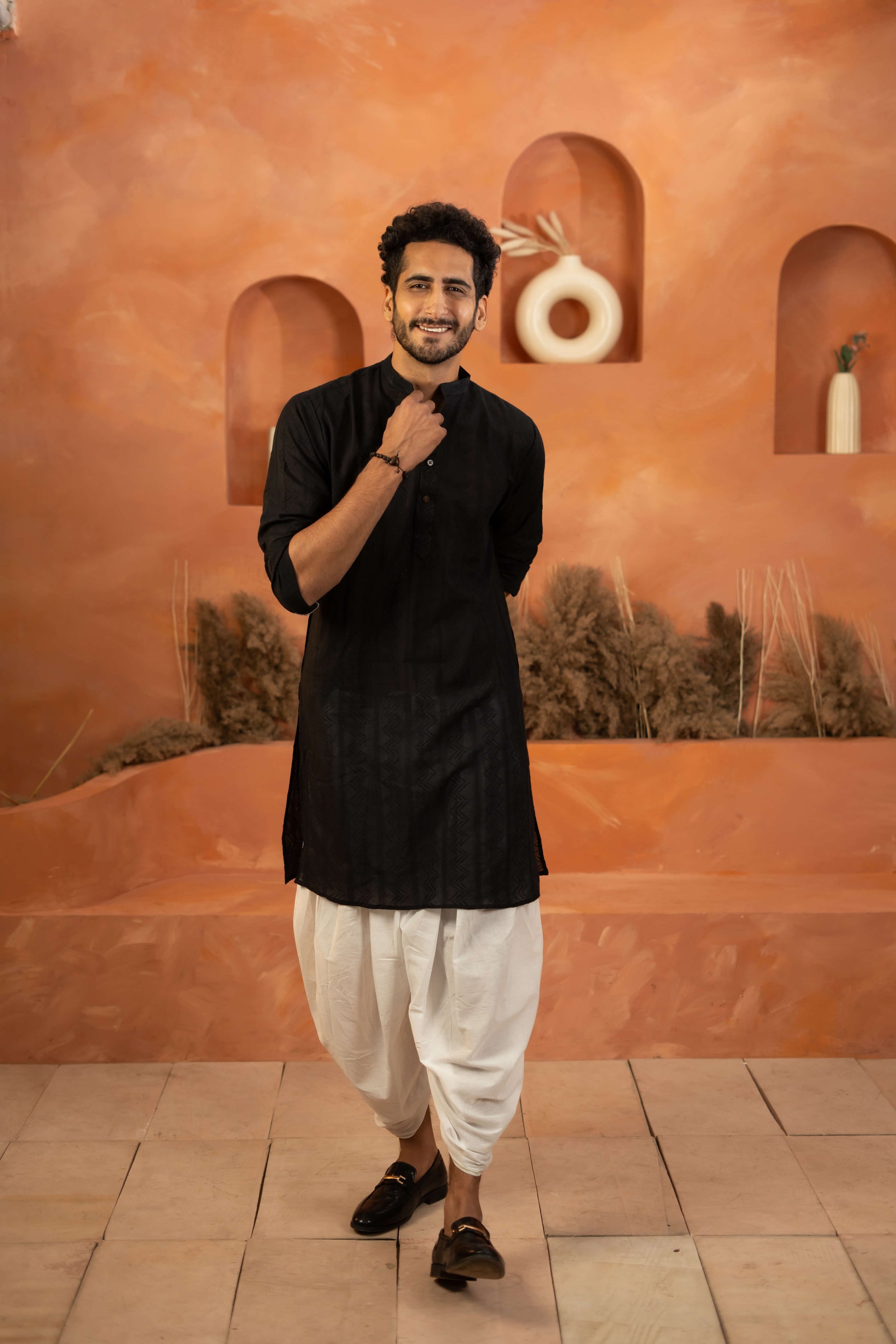 Person wearing a black kurta and white trousers, standing by an orange background.