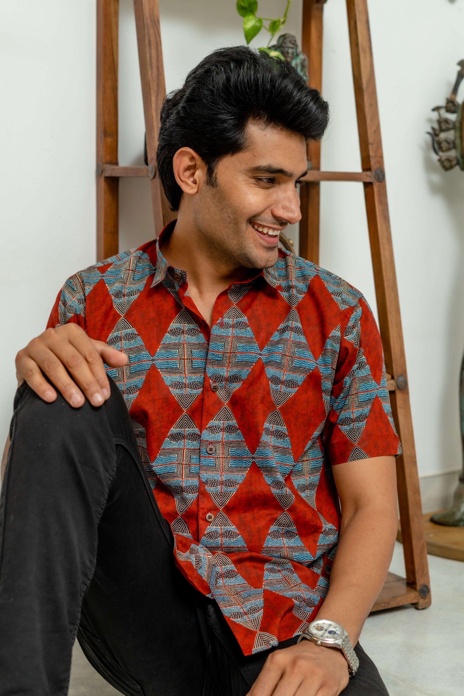 Indian man featuring a red color half sleeve shirt. this shirt featuring beautiful diamond print. this man is wearing a analog watch sitting in front of a wooden stand