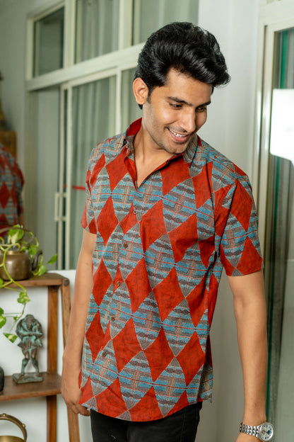 Indian man in red color shirt featuring diamon print. this man is wearing a analog whatch and standing in front of a wooden stand.