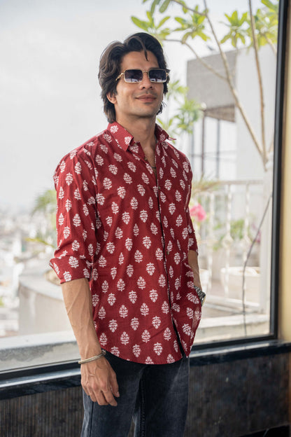 Indian man wearing a red printed shirt featuring a white floral print. Wearing sunglasses and standing casually in front of a mirrored wall. 