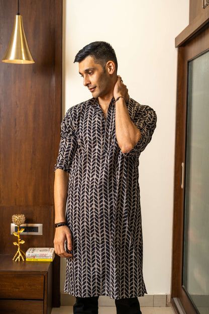 Indian man wearing a black color long kurta featuring beautiful petal print. standing in front of a beautiful wall decorated with wooden interior decorated with some books and golden color lamp