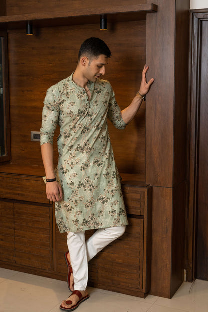 Man wearing a pastel green floral kurta with white trousers and brown sandals, posing stylishly against a wooden interior background.