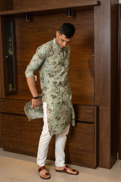 Man in a pastel green floral kurta paired with white trousers and brown sandals, adjusting the hem of the kurta while standing against a wooden interior backdrop.